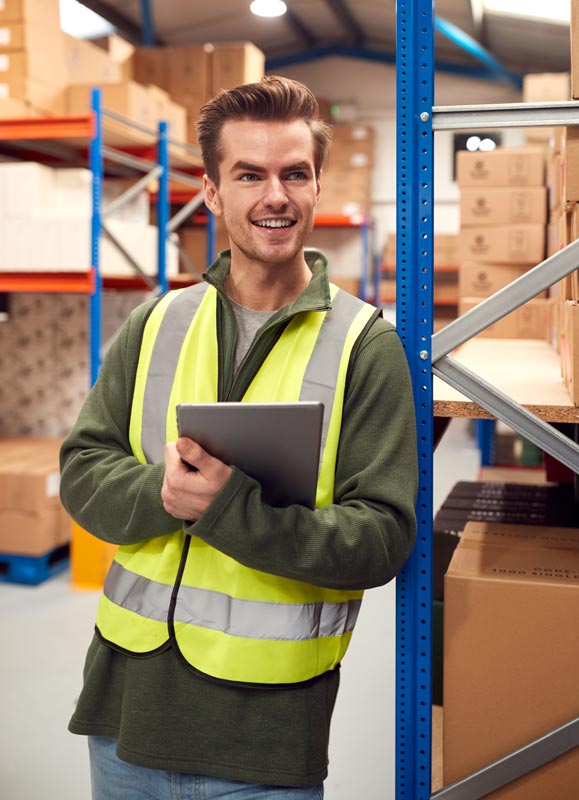 A male worker inside the warehouse checks stock on shelves using digital tablet.